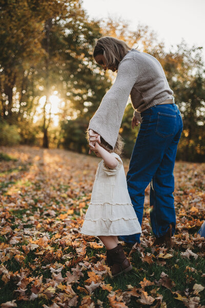A mother and her young daughter share a tender moment, holding hands while walking through autumn leaves at sunset. Captured during a fall family photography session in Connecticut by Nicole Mele Photography, celebrating love and connection in nature.