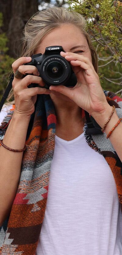 Blonde haired girl outside holding camera to eye, taking photo straight on. She's wearing a multi-color tribal scarf and a white shirt.