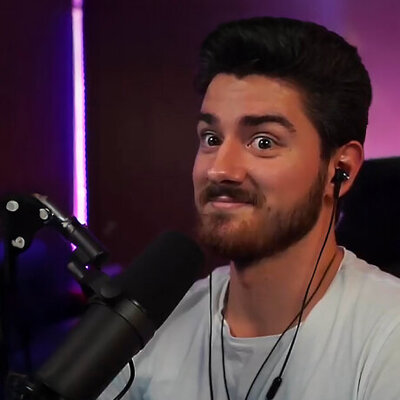 Man sitting in a purple-lit room with short brown hair and full beard and mustache. He is smiling slightly and wearing a white shirt. A microphone is propped next to his mouth.