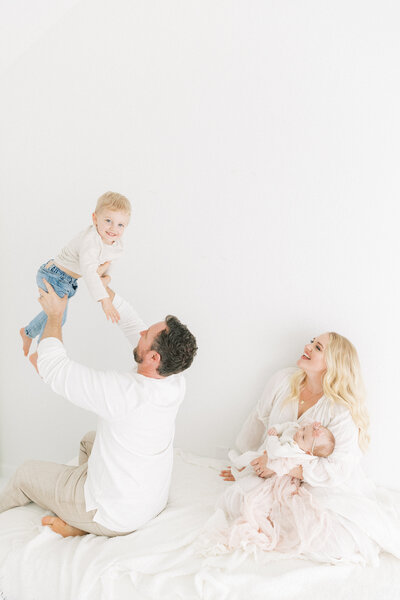 Mom sitting in white dress in photography studio holding newborn baby girl while dad holds older brother up in the air smiling