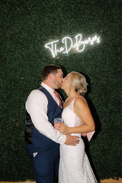Bride and groom kiss in front of a grass wall with their last name illuminated behind them in neon