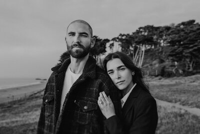 Black and white portrait of a couple standing by the coast, with the woman leaning into her partner. Their expressions are calm and introspective, framed by trees and ocean in the background.