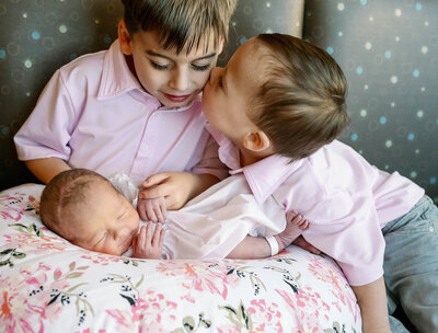 Two older brothers wearing pink shirts holding their newborn baby sister on a pillow.