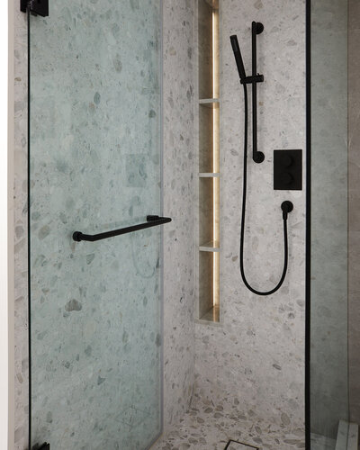 A walk-in shower featuring elegant stonework walls and flooring, built-in, backlit shelving, a black showerhead fixture, and a hinging glass door
