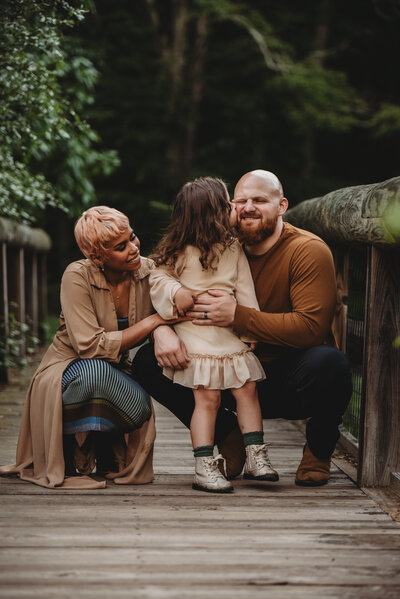 A young girl lovingly kisses her father’s cheek while her mother smiles, capturing a heartfelt family moment on a rustic bridge during a lifestyle family photography session in Connecticut. Captured by Nicole Mele Photography, highlighting love, connection, and joy.