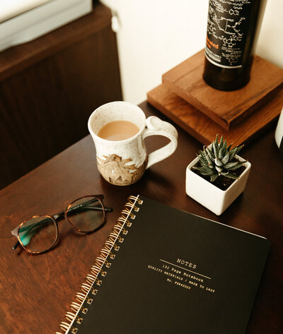 A cup of coffee, pair of glasses, notebook and plant sitting on desk