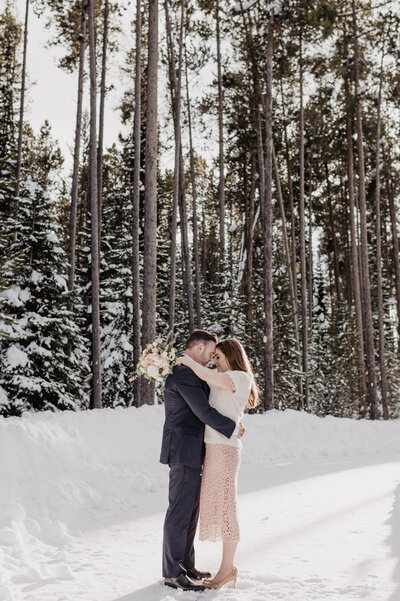 winter elopement captured by Jackson Hole photographers with groom holding bride around the waist as she holds his shoulders as they stand on a path in front of pine woods covered in snow