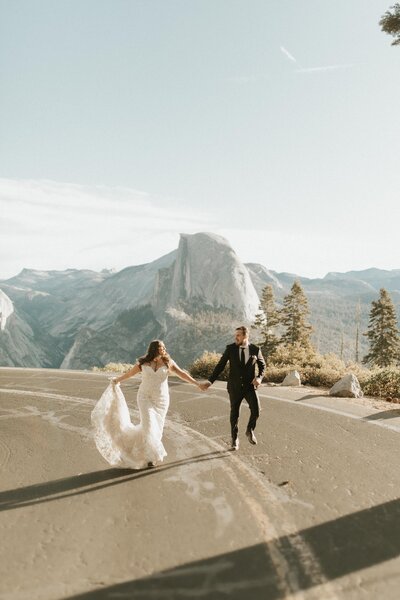 man and woman holding hands and running together in the middle of the road with a mountain backdrop