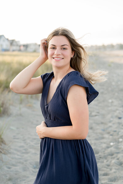 girl standing on the beach wearing a blue dress, touching her hair and smiling