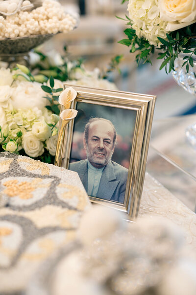 A framed portrait of an older man with a beard, wearing a suit, is decorated with white calla lilies and placed on a table surrounded by flowers.