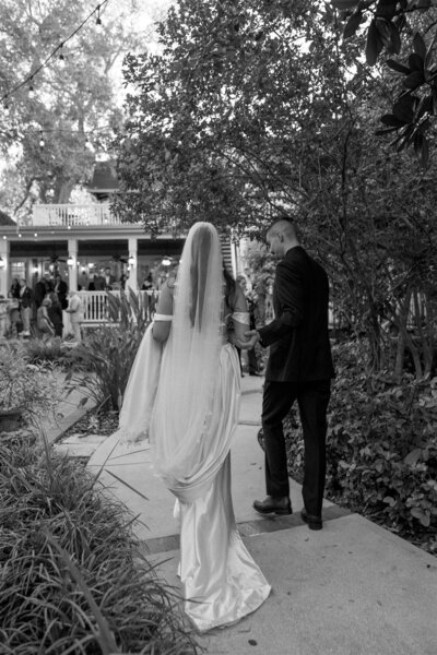Black and white wedding photo of a bride in an off-the-shoulder satin gown and veil walking hand-in-hand with her groom through the garden pathway at Dr. Phillips House in Downtown Orlando, Florida, with guests gathered on the porch in the background.