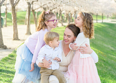 Salt Lake Photography of a mother, her two young daughters, and her toddler son smiling and laughing at each other with blossoming cherry trees in the background.