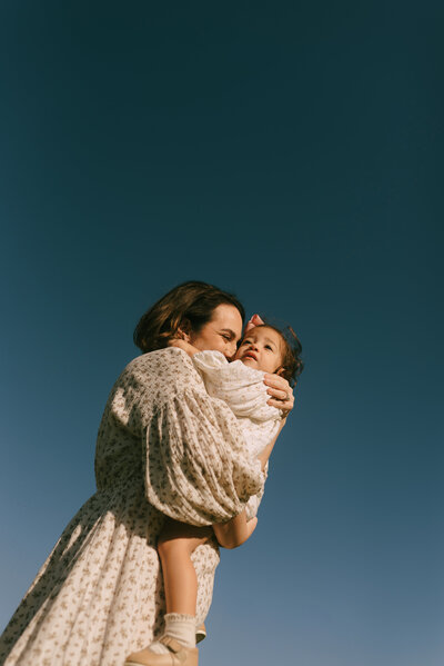 A mother holding her daughter, cuddling her close, with the deep blue sky as their background. 