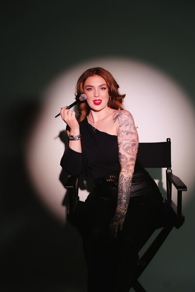 MaKall seated in a director’s chair holding a makeup brush, looking confidently at camera