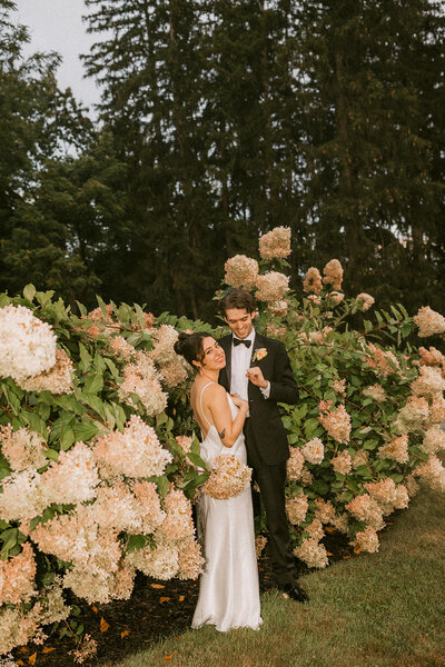 A joyful couple poses amidst blooming hydrangeas on their wedding day, captured by a timeless natural light wedding photographer in a romantic outdoor setting.
