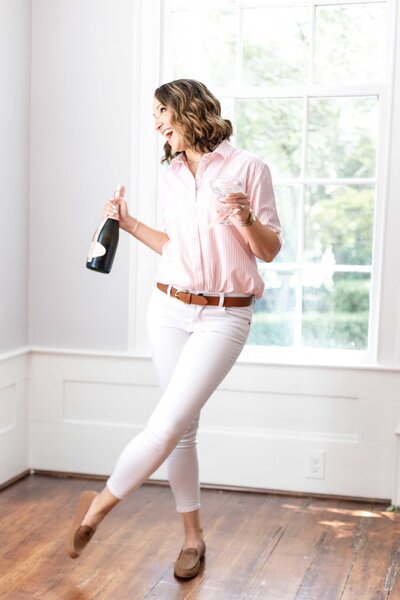 A luxury party rentals expert dances with a champagne bottle in hand, dressed in a white pant and pink blouse. The scene captures the joy and celebration associated with high-end event services.