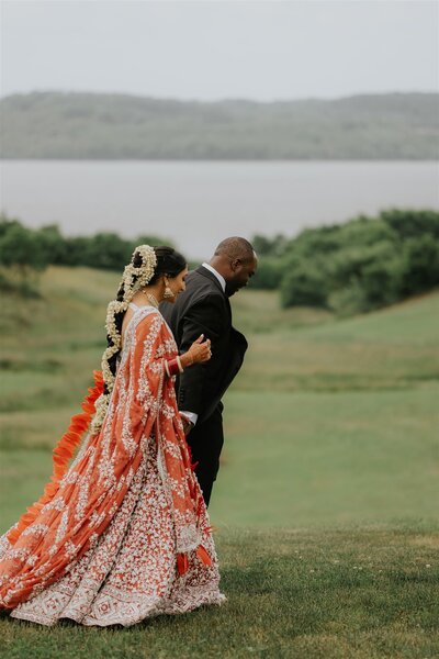 Bride and Groom walk along rolling grassy hills overlooking a lake and Hudson Valley mountains. Bride wears traditional Indian wedding gown.