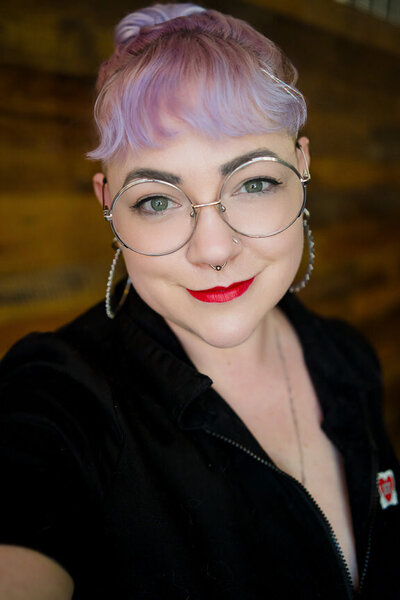 A person with purple hair and large, round glasses smiling while standing in front of a wood wall.