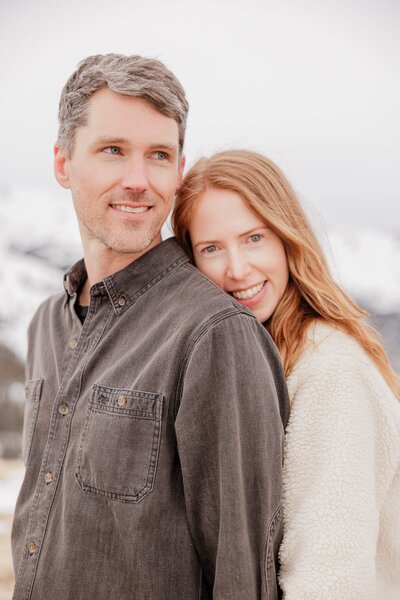 A laughing man gives his partner a piggy back ride in blue shirts in snow during their Breckenridge Couples Photography session