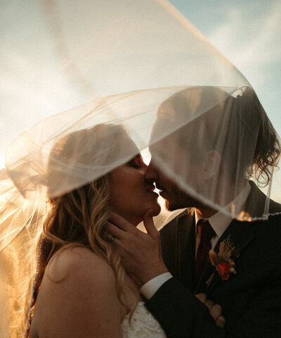 Bride and groom kissing under veil at sunset