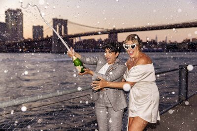A couple standing on the edge of the water popping a bottle of champagne.