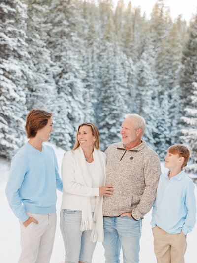 A happy family of 4 with 2 teen boys laughs while standing in a snowy mountain pass