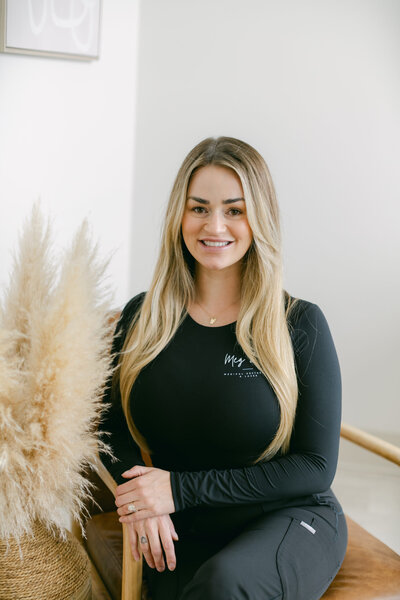 A woman with blonde hair wearing black scrubs sits smiling on a brown chair in a bright room at Falmouth Maine Medical Aesthetics, with pampas grass visible to the side.