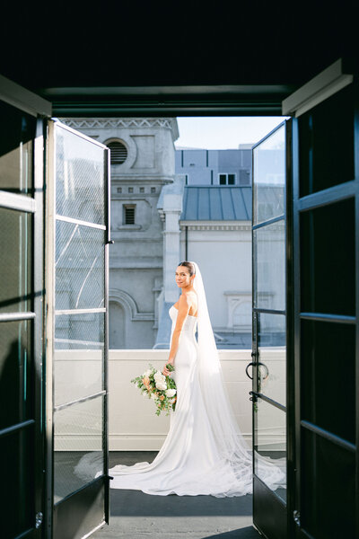 bride stands on the balcony at Vibiana and her portrait is taken through the doors overlooking the city of los angeles captured by los angeles wedding photographer magnolia west photography