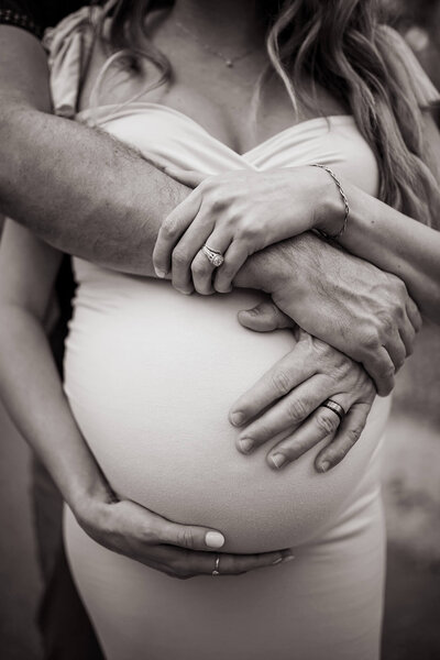 Black and white detailed portraiture of man and woman's arms interlinked around pregnant belly 