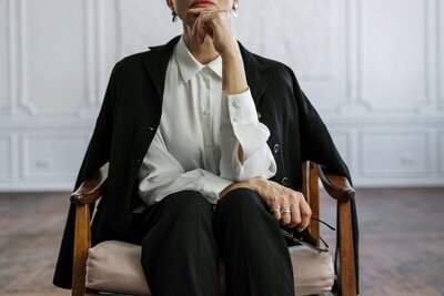 A reflective woman seated in a wooden armchair, dressed in a white blouse and black blazer, resting her chin on her hand in a poised and contemplative pose.