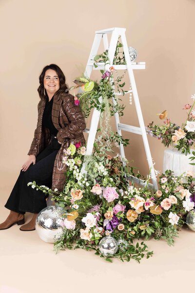 Emily Rey leans on a ladder adorned with flowers. Representing her vendor profile for the Crush Collective wedding show.