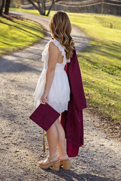 High school senior in cap and gown near gravel road with golden light filtering through trees