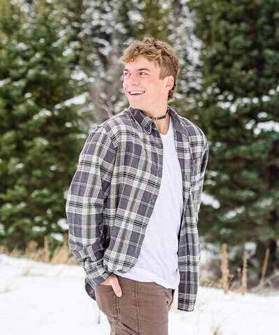 salt lake city senior photography of  high school senior boy wearing a gray plaid shirtl ooks over his shoulder as he stands in the snow at Tibble Fork Canyon in American Fork