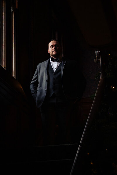 A groom standing on a dark staircase illuminated by window light at lord thompson manor