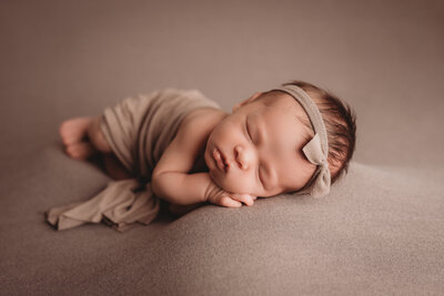 Newborn baby girl asleep at Marietta, GA newborn portrait studio posing on side wearing taupe wrap and taupe bow headband.
