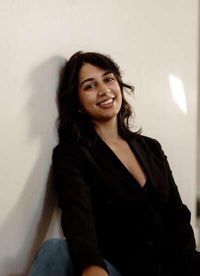 A person with shoulder-length dark hair smiles while leaning against a wall. They are wearing a black blazer and jeans, sitting casually with a relaxed posture. The lighting creates soft shadows on the wall.
