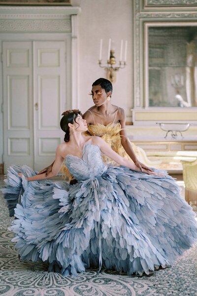 Two models in elegant Karen Sabag gowns stand in a luxurious room in villa Pizzo, Lake Como, captured by  international wedding photographer Andreas K. Georgiou. One wears a voluminous blue dress adorned with feathers, while the other dons a flowing yellow gown. Gracefully gazing at each other amid ornate classical decor, their pose is timeless.