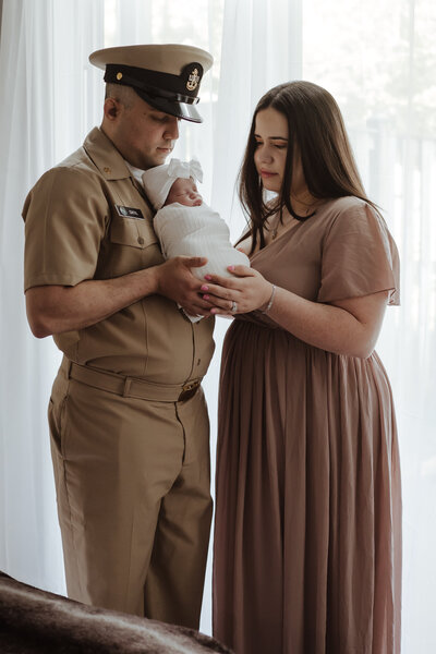 Dad dressed in US Navy khaki uniform holding baby girl wrapped in white while mom in neutral dress is holding bottom of baby while looking at baby in Severn, Maryland studio photographed by Bethany Simms Photography