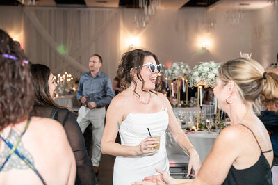 A bride in heart-shaped sunglasses laughs and dances with guests during the reception at SoHo63, a Chandler wedding and event venue. Holding a cocktail and surrounded by floral centerpieces and glowing decor, she celebrates in a lighthearted, joyful atmosphere.