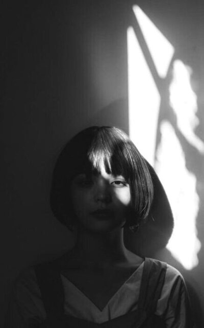 Black and White Portrait of a woman with a fringe and short bob hairstyle against a wall with shadows and  light reflecting from a window.