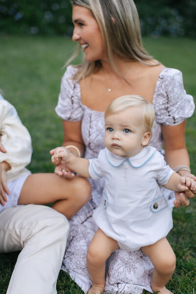 Mom sitting in the grass holding her baby's hands as he stands in front of her