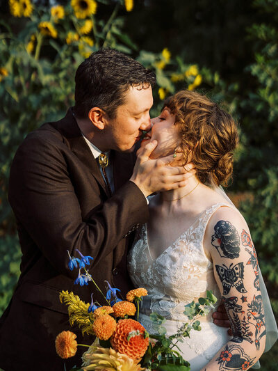A bride and groom kissing as she holds her bouquet and he touches her face. 