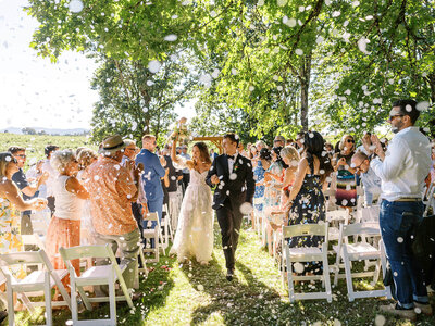 A bride and groom walk down the aisle at an outdoor wedding while guests clap and throw flower petals.