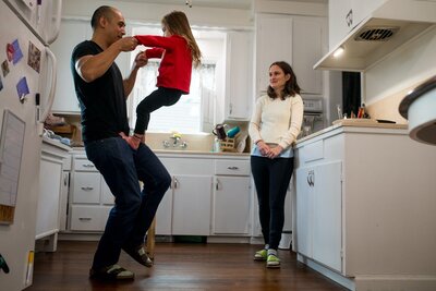 A little girl wearing a red sweater holds her dads hands and climbs up his legs in their kitchen while mom watches. 