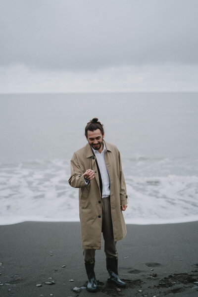 Ein Mann im Trenchcoat und Gummistiefeln lächelnd am Strand. Hinter ihm die Gischt – bereit für Abenteuer und positive Veränderungen