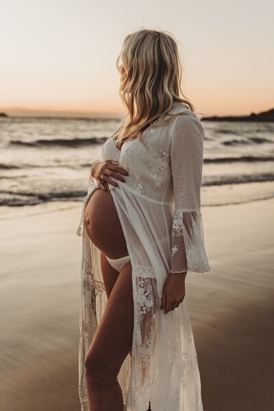 A pregnant woman in a sheer white dress stands on a beach at sunset, holding her belly and facing away from the camera, perfectly capturing the serenity of Wollongong motherhood sessions by NSW family photographer.