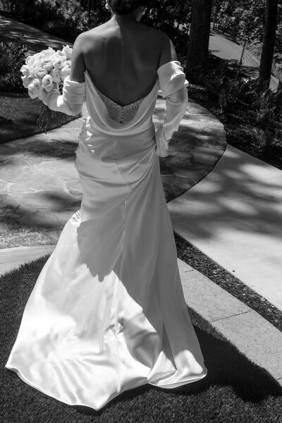 Rear view of a bride wearing a fitted satin wedding dress with a draped cowl back, holding a bouquet while walking on a stone path.