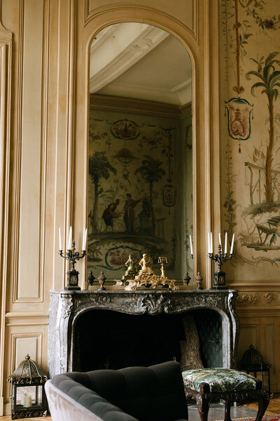 An elegant French chateau interior featuring an ornate fireplace with detailed candelabras, gold accents, and classic European decor