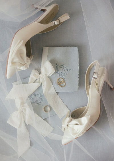 Detail shot of a brides rings, shoes, and vow books on a light blue backdrop