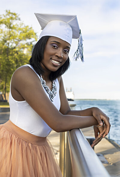 Senior girl with long peach tutu skirt, white sleeveless top, and white graduation cap leaning on rail overlooking the Detroit River